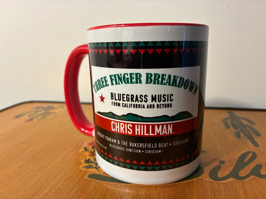 A ceramic mug with a red handle and red rim, featuring a printed label for Chris Hillman's 'Three Finger Breakdown' bluegrass music. The mug is resting on a wooden table with a white wall in the background.
